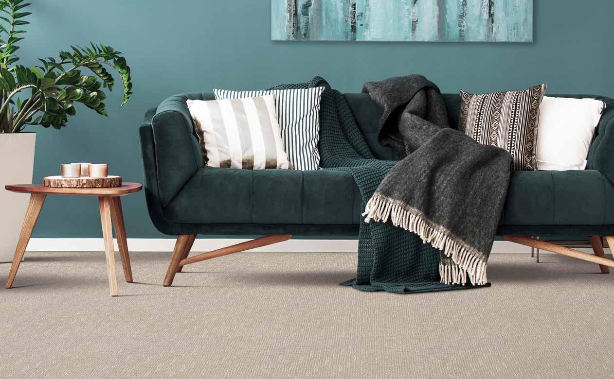 Modern living room with teal walls and abstract blue toned art, with a light grey patterned carpet, dark teal sofa, patterned pillows, wooden side table with candles, and a tall green plant.